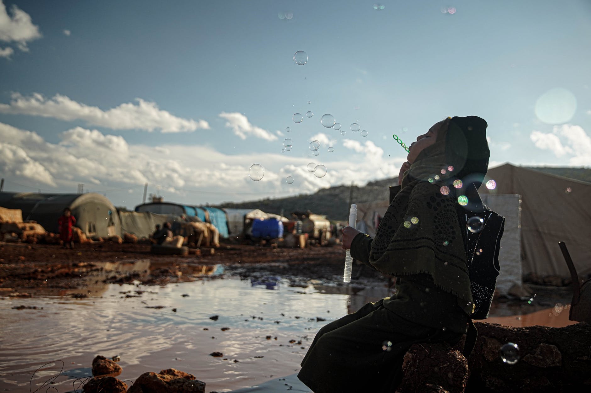 hijabi woman kneeling by puddle in refugee camp blowing bubbles